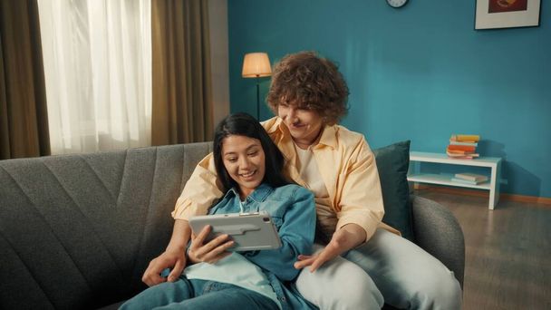 A young married couple spends time together on a day off. a Woman and a man watch a movie, photo, video on a tablet. A young Asian woman lies on a sofa with a tablet in her hands, leaning her back on - Foto, imagen