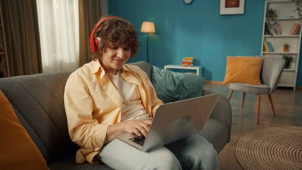 A young man is sitting on a couch wearing red wireless headphones with a laptop on his lap. A man is typing a text, a message, chatting, listening to music, playing online games. Close up. - Foto, Imagem