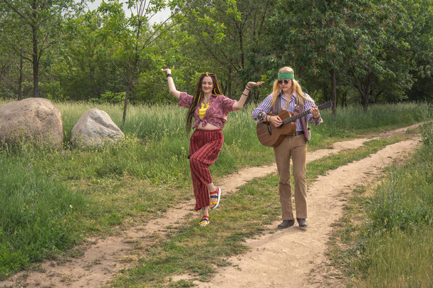 Couple of young people in hippie style. A girl is dancing, a guy is playing a guitar on a country road on a sunny day - Фото, зображення