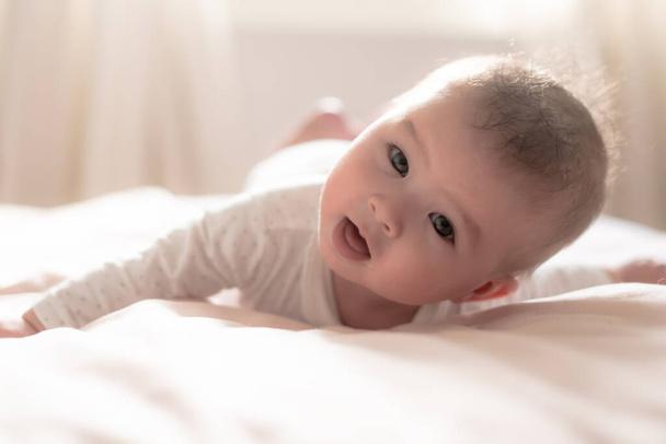 adorable y feliz niña de 5 meses descubre alegremente alegría ilimitada mientras explora juguetonamente su cama. Rodeado por el cálido resplandor de la luz natural, el pequeño sonríe alegremente - Foto, Imagen