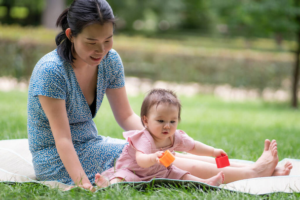 outdoors lifestyle portrait of mother and daughter - young happy and sweet Asian Korean woman playing with her 8 months baby girl on grass at city park - Photo, image