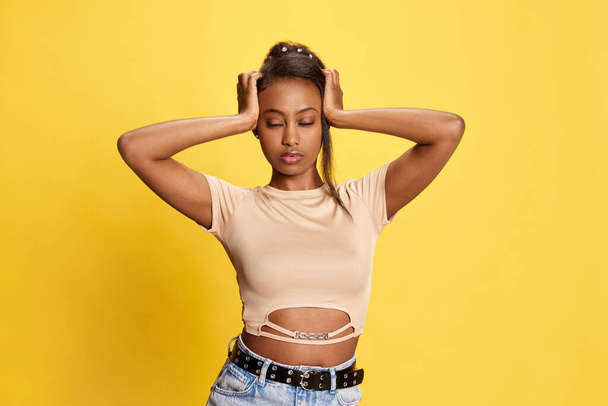 Portrait of pretty young african girl holding hands on head with thoughtful, concentrated face against yellow studio background. Concept of lifestyle, human emotions, fashion, beauty, ad - Foto, Imagem