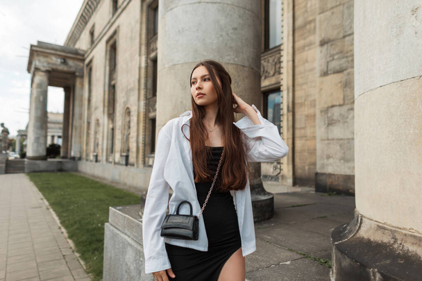 Beautiful young stylish chic girl model in a fashion black dress with a white shirt and a leather handbag walks in the city near a vintage building with columns - Photo, Image
