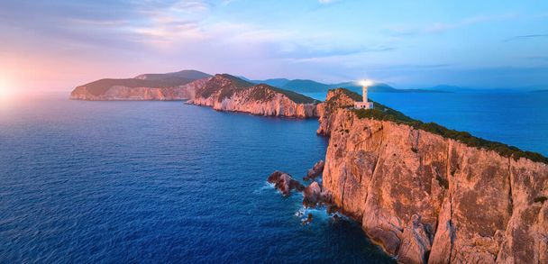 Panoramic, aerial view of Cape Ducato cliffs and sea with lighthouse shining, illuminated by pink light of setting sun. Lefkada, Greece. - Photo, Image
