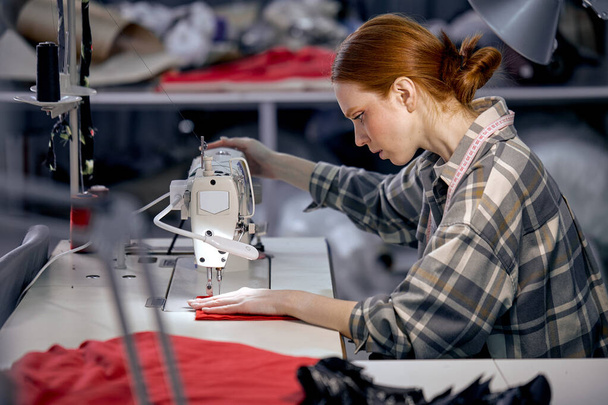 seamstress lady in casual wear sit behind table using sewing machine, making modern clothes, young redhead caucasian woman at workshop, holding red fabric, enjoy tailoring, controlling equipment - Photo, Image