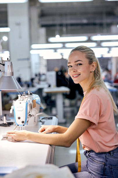 young nice caucaian blonde textile factory worker on production line using modern sewing machine, at workshop. hardworking woman engaged in sewing or tailoring,looking at camera smiling - Foto, immagini