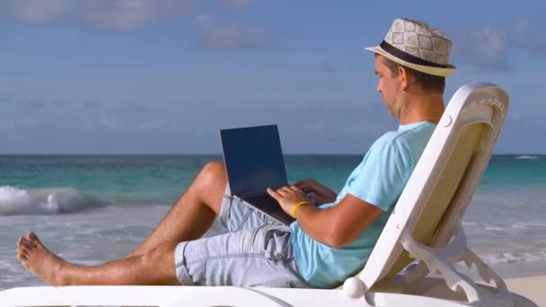 Freelance à la plage. Mode de vie jeune homme caucasien travaillant sur ordinateur portable tout en étant assis se détendre sur la belle plage tropicale en plein air, travailleur indépendant social pendant les vacances d'été. Concept d'été et vacances - Séquence, vidéo