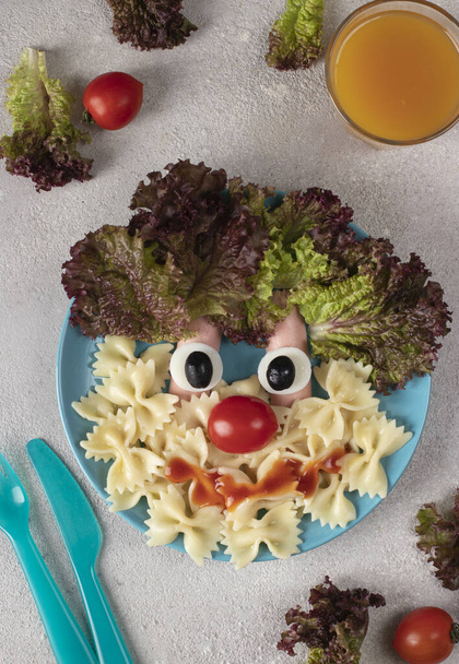 Fun Food for kids - smiling face from pasta, sausages, egg, cherry tomato and lettuce leaves on blue plate, Top view - Foto, afbeelding
