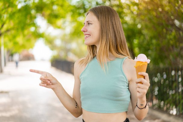 Молодая блондинка красивая женщина с мороженым из корнета на открытом воздухе, указывая в сторону, чтобы представить продукт - Фото, изображение