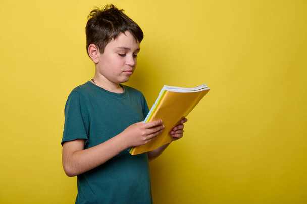 Adorable Caucasian schoolboy in green casual t-shirt, primary school student, holds colorful textbooks, isolated on yellow background. Back to school. Education concept. Smart clever kids - Photo, Image