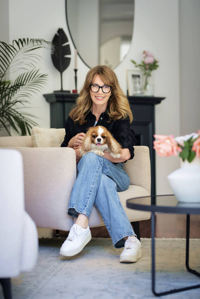Happy attractive woman relaxing in an armchair at home. Mid aged female sitting at home with her cute cavalier king charles puppy. - Фото, зображення