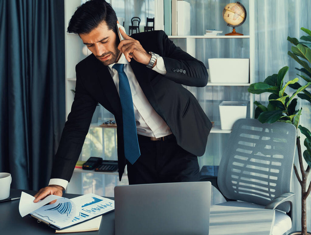 Een ijverige zakenman die in gesprek is met klanten terwijl hij met een laptop in zijn kantoor werkt als concept van een moderne hardwerkende kantoormedewerker met mobiele telefoon. Vurig - Foto, afbeelding