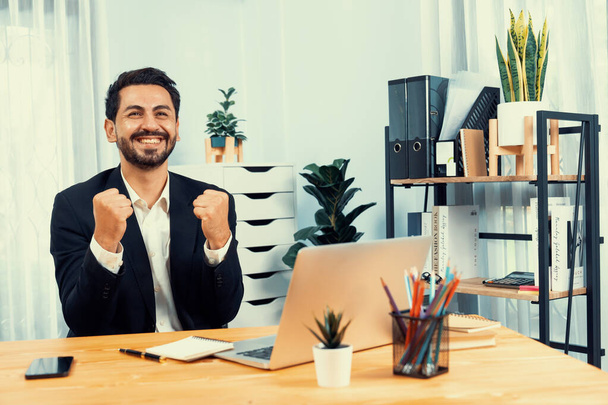 Excited and happy businessman dressed in black formal suit, raise his arm in celebratory gesture at his office desk, after successful job promotion, energy and joy as business winner. Fervent - 写真・画像