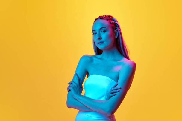 Portrait of beautiful, attractive young girl with cute hairstyle looking at camera with calm face against yellow studio background in neon light. Concept of youth, emotions, beauty, lifestyle, ad - Valokuva, kuva