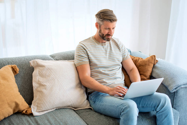 Middle aged man sitting on sofa at home and usng laptop for work, video call. Caucasian male wearing casual clothes. - Foto, Imagem