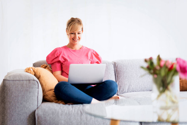 Blond haired woman sitting on sofa and using laptop for work or having video call. Confident female wearing casual clothes and smiling. Home office.  - Fotografie, Obrázek