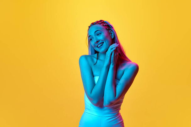 Happy, caring look. Portrait of attractive young woman with pretty, stylish hairstyle posing against yellow studio background in neon light. Concept of youth, emotions, beauty, lifestyle, ad - Φωτογραφία, εικόνα