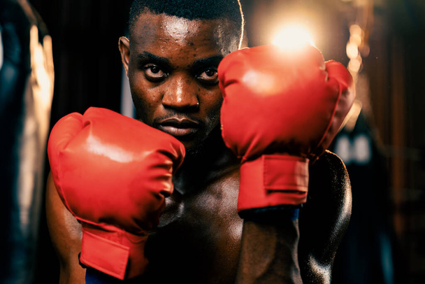 Boxkämpfer in hemdsloser Pose, afroamerikanischer schwarzer Boxer mit rotem Handschuh in defensiver Wachhaltung, bereit zum Kampf und zum Schlagabtausch im Fitnessstudio mit Kickbag und Boxausrüstung im Hintergrund. Impulse - Foto, Bild