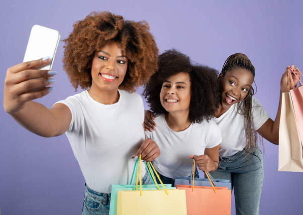 Mobiel winkelen Plezier. Drie vrolijke Afro-Amerikaanse vrouwen kopers maken selfie op telefoon, poseren met papieren shopper tassen staande over Purple Studio achtergrond. Groot e-commerce aanbod - Foto, afbeelding