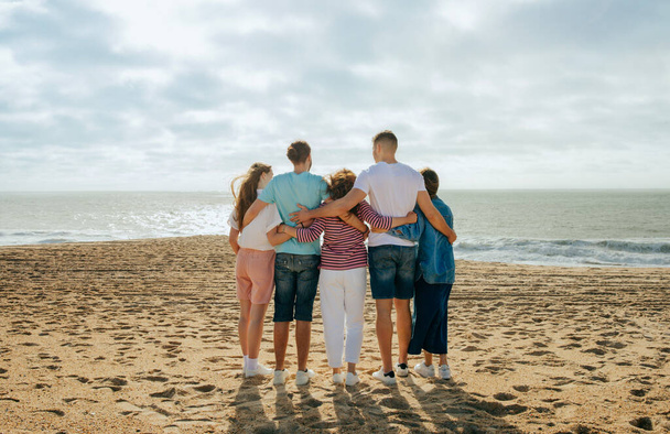 Millennial european and arab people in casual hugging, enjoy freedom, team building on beach at weekend, outdoor, back. Free time with friends, holidays and walk together in summer, ad and offer - Photo, Image