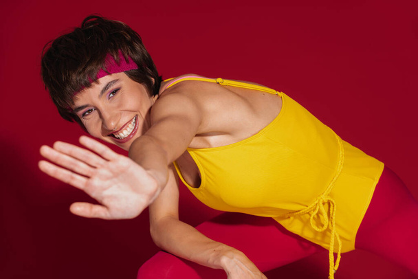 Happy young fit woman in retro styled sportswear stretching out hand against red background - Fotó, kép