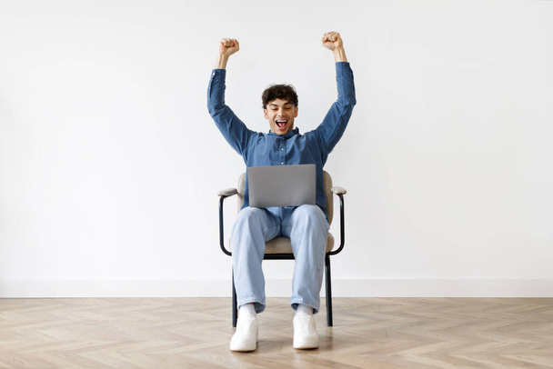 Emocionado hombre árabe agitando puños usando el ordenador portátil, mirando a la cámara y celebrando el éxito empresarial o grandes noticias en línea, posando Agaisnt pared blanca en casa, sentado en una silla de ruedas - Foto, Imagen