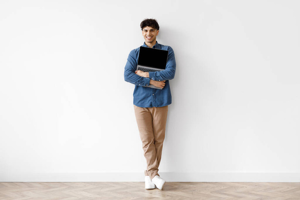 Embracing Online Technology. Full Length Of Happy Arab Guy Holding And Hugging Laptop, Advertising Internet Offer, Leaning On White Wall Standing Indoor. Technology And Freelance, Copy Space - Foto, imagen