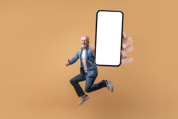 E-commerce, retail, great offer. Excited grey-haired senior man jumping in the air with huge smartphone with white blank screen in his hand, beige background, showing thumb up, mockup, copy space - Photo, Image