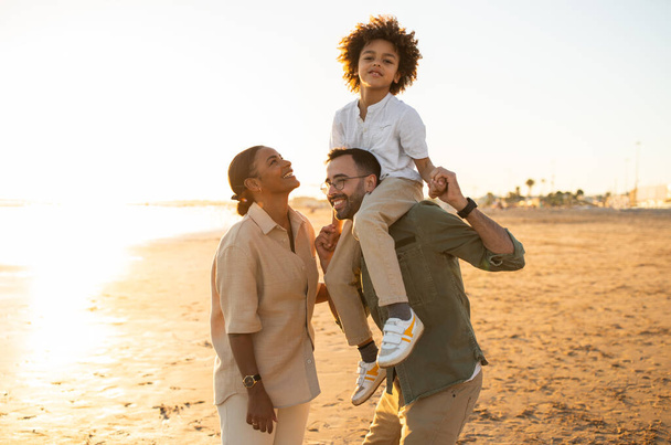 Joyeux jeune famille noire de trois personnes profitant du temps ensemble au bord de la mer à l'extérieur, père tenant son fils sur les épaules, marchant et s'amusant le soir - Photo, image
