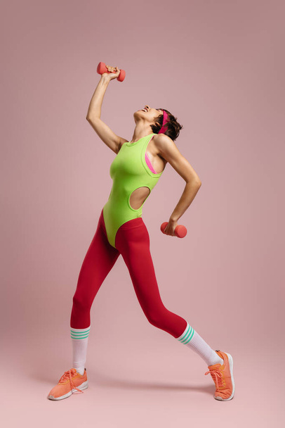 Happy fit woman in retro styled sports clothing exercising with dumbbells against colored background - Φωτογραφία, εικόνα
