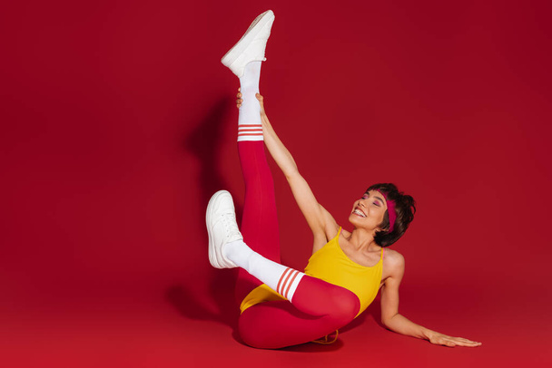 Playful young fit woman in retro styled sports clothing stretching against red background - Foto, Imagen
