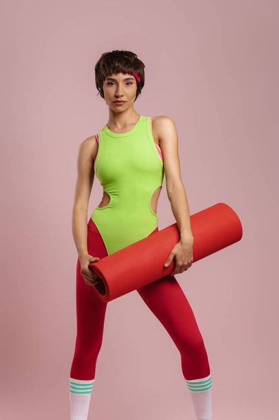 Beautiful fit woman in retro styled sports clothing carrying exercise mat against colored background - Fotó, kép