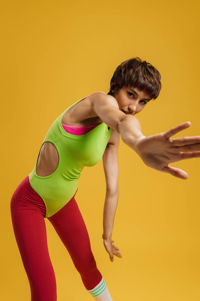 Young fit woman in retro styled sports clothing stretching out hand against yellow background - Foto, afbeelding