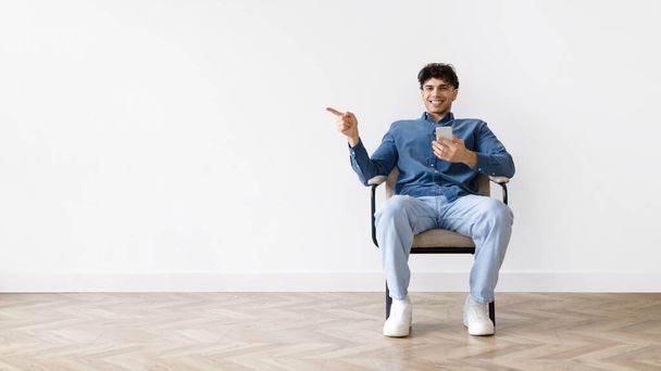 Mobile Application. Positive Young Arab Guy With Cellphone Pointing Finger Aside At Empty Space, Advertising App Offer, Sitting In Armchair Against White Studio Wall Indoor. Panorama - Photo, Image