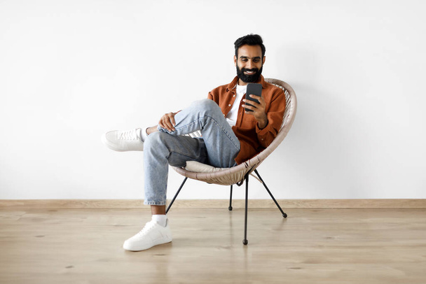 Application mobile. Homme Moyen-Orient joyeux textos sur Smartphone assis dans le fauteuil à confort de la maison, posant sur fond blanc. Guy utilisant une nouvelle application sur le téléphone jouissant week-end confortable - Photo, image