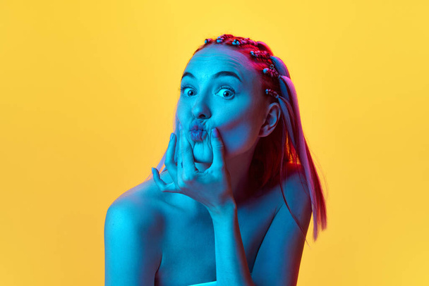Portrait of cute, pretty young girl with stylish hairstyle posing with grimace face against yellow studio background in neon light. Fun and joy. Concept of youth, emotions, beauty, lifestyle, ad - Valokuva, kuva