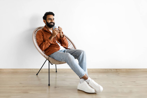 Young arabic guy with cup of hot coffee chilling in cozy armchair against white studio wall indoor, looking aside at empty space. Relaxed millennial man having break relaxing at home - Photo, Image