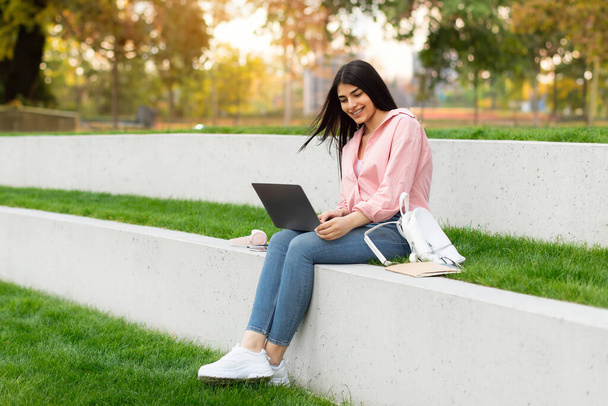 Distance education concept. Happy hispanic student lady with laptop sitting outdoors and watching educational webinar, looking at screen and smiling, copy space - Φωτογραφία, εικόνα
