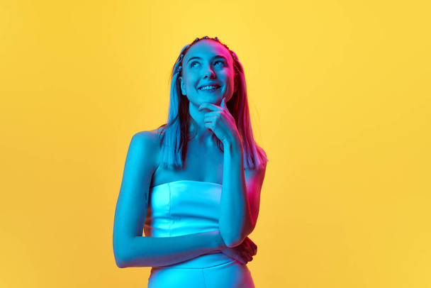 Portrait of beautiful young woman with cute hairstyle standing with smile and dreamy, thoughtful look against yellow studio background in neon light. Concept of youth, emotions, beauty, lifestyle, ad - Zdjęcie, obraz