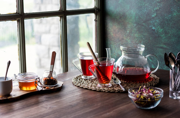 Υγιές τσάι ιβίσκου σερβίρεται με μέλι σε ένα σκοτεινό τραπέζι κοντά σε ένα παράθυρο.  - Φωτογραφία, εικόνα