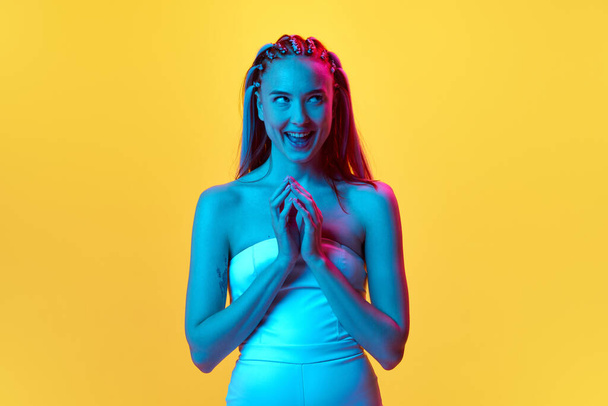 Making joyful plan. Portrait of pretty young woman with funny thoughtful face standing against yellow studio background in neon light. Concept of youth, emotions, beauty, lifestyle, ad - Zdjęcie, obraz