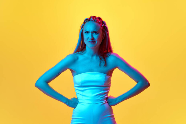 Portrait fo young emotive girl standing with angry, unpleasant face and looking attentively at camera against yellow studio background in neon light. Concept of youth, emotions, beauty, lifestyle, ad - Foto, Imagem