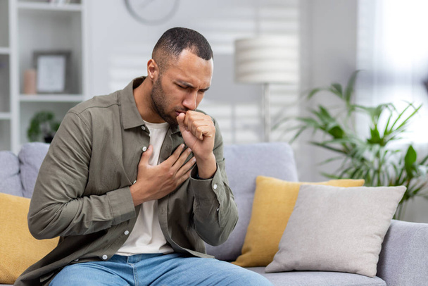 Young African American man sitting on the couch at home and coughing. Suffers from an attack of asthma, allergies. He holds his chest, covers his mouth with his hand. - Foto, Imagen