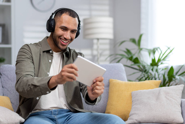 A young Hispanic man plays online games on a tablet at home. Sitting smiling on the couch wearing headphones. - 写真・画像