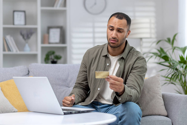 Upset young hispanic man using credit card and laptop at home on sofa. Unsuccessful online shopping, hacked account, bankruptcy, fraud. - Photo, image