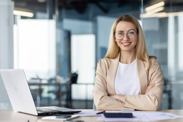 Portrait of a successful and self-confident business woman in a business suit, sitting in the office at the desk and looking at the camera, smiling with her arms crossed. - Photo, image