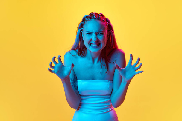 Portrait of young emotive girl standing with extremely angry expression against yellow studio background in neon light. Irritation. Concept of youth, emotions, beauty, lifestyle, ad - Foto, immagini