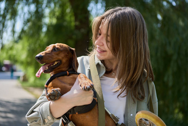 Pet owner carrying a paralyzed dachshund on a wheel chair. Cheerful young girl holding a handicapped dog in hands during a walk outdoor - Fotoğraf, Görsel