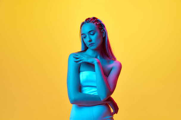 Portrait of pretty, young girl with cute hairstyle, in white clothes posing with sad expression against yellow studio background in neon light. Concept of youth, emotions, beauty, lifestyle, ad - Фото, изображение
