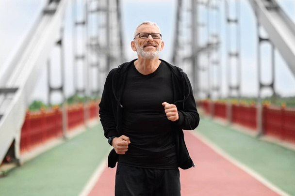 Cheerful handsome grey-haired athletic senior man wearing black sportswear running outdoors by bridge in the morning. Mature sportsman enjoying jogging by street. Healthy lifestyle for older adults - Photo, Image